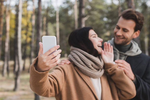 excited woman taking selfie on cellphone with boyfriend in forest on blurred background - Foto, imagen