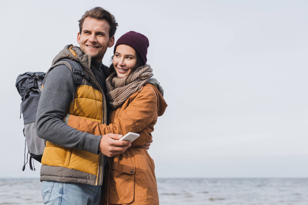 cheerful man in autumn outfit holding smartphone while hugging with girlfriend near sea - Fotoğraf, Görsel