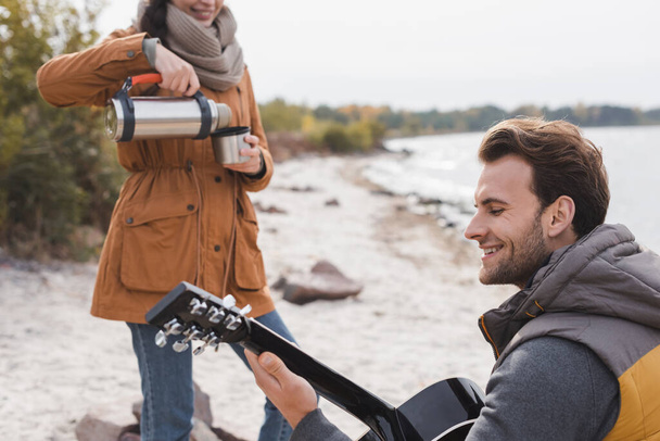 cheerful man playing acoustic guitar while woman pouring drink from thermos during autumn walk - 写真・画像