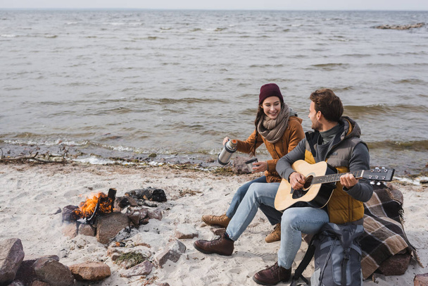 man playing guitar to happy woman with thermos while sitting near bonfire on sea coast - 写真・画像