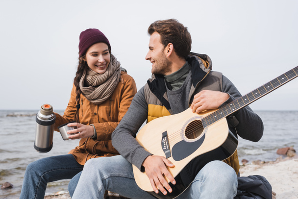 秋の散歩中にギターを持って川沿いに座っているテルモスと男を持つ陽気な女性 - 写真・画像
