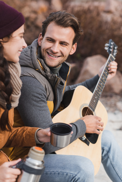 счастливый мужчина играет на акустической гитаре размытой женщине с термосом  - Фото, изображение