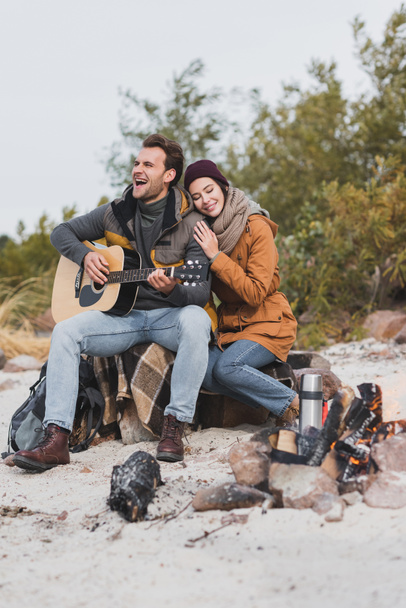 happy woman with closed eyes leaning on man playing guitar during halt in autumn walk - Photo, Image
