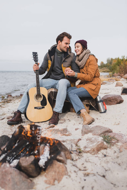 happy woman and man with guitar sitting near blurred bonfire at sea coast - Foto, imagen