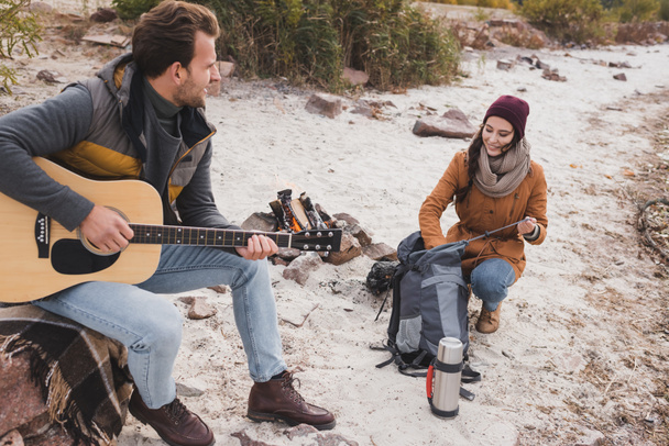 man playing guitar to smiling woman near backpack and thermos - Fotoğraf, Görsel