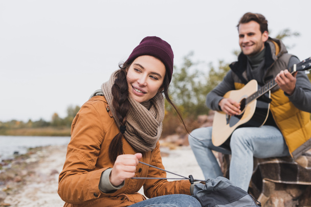 cheerful woman looking away and holding backpack near blurred man playing acoustic guitar - Photo, Image