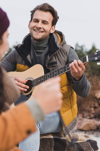 happy young man in autumn outfit playing guitar to blurred woman during autumn walk - Photo, Image