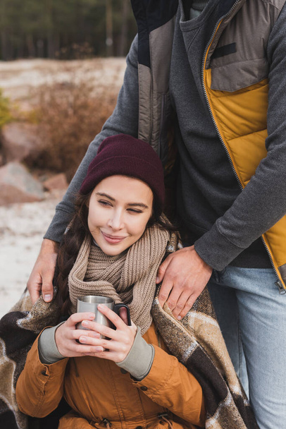 smiling woman holding thermo cup near man covering her with plaid blanket outdoors - Photo, Image
