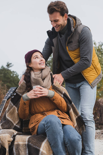 young man in autumn clothes covering girlfriend with warm blanket during halt in walk - Photo, Image