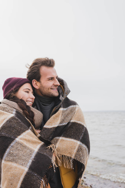 cheerful couple, wrapped in warm plaid blanket, looking away near sea - Foto, Imagen