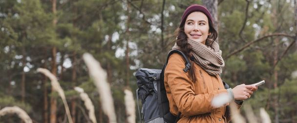 mujer positiva con mochila y teléfono móvil senderismo en el bosque en primer plano borrosa, bandera - Foto, Imagen