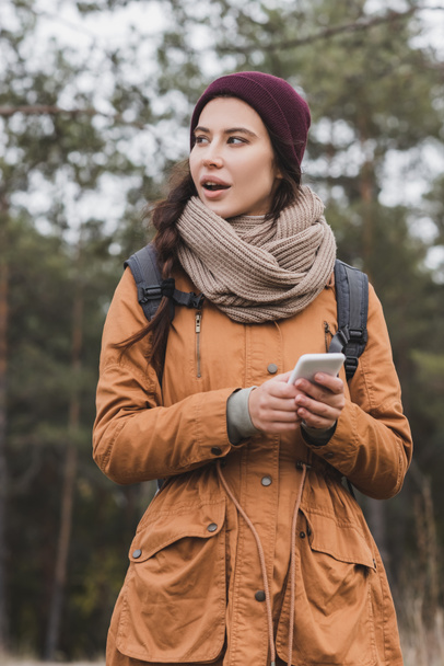 thoughtful woman with cellphone looking away while hiking in forest - Photo, Image