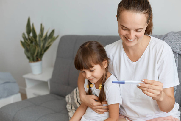 Caucasian young adult woman wearing white t shirt holding pregnancy test in hands, sitting on sofa at home with her daughter, rejoicing positive result. - Fotografie, Obrázek