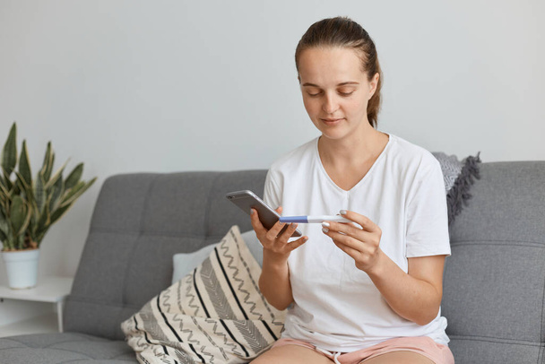 Optimistic pregnant Caucasian dark haired female wearing casual style clothing with pregnancy test in hands, and holding smart phone in hands, dialing husband number to tell him good news. - 写真・画像