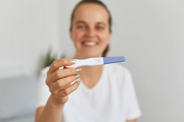 Indoor blurred shot of smiling young attractive female with pigtail holding out pregnancy test, sitting on bed in bedroom, being happy to be future mother. - Foto, Bild