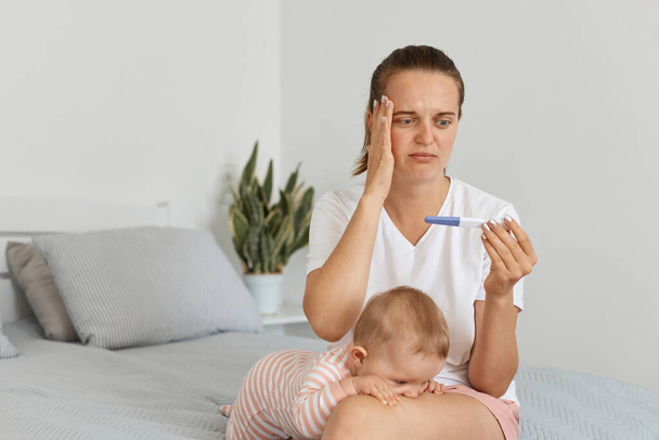 Indoor shot of sad young adult pregnant woman with pregnancy test, posing in her bedroom with toddler baby, wearing white t shirt, touching her head, having serious problems. - Foto, afbeelding