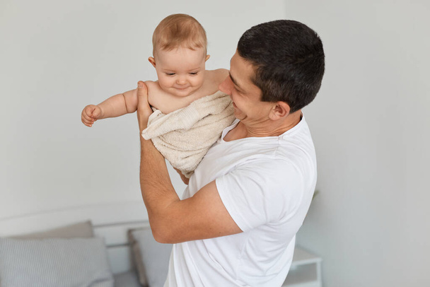 Portrait of handsome optimistic young adult father with his toddler baby girl, looking at kid wrapping in towel after bathroom, playing together at home. - Foto, Imagem