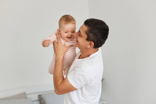 Side view portrait of loving young adult father with dark hair, holding his toddler baby, girl playing together at home, happy parenthood and childhood. - Foto, Imagen