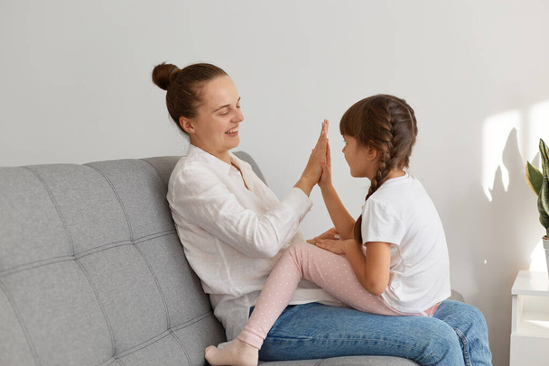 Side view portrait of mother and her daughter playing patty-cake while sitting on sofa, family posing indoor in living room, motherhood and childhood. - 写真・画像