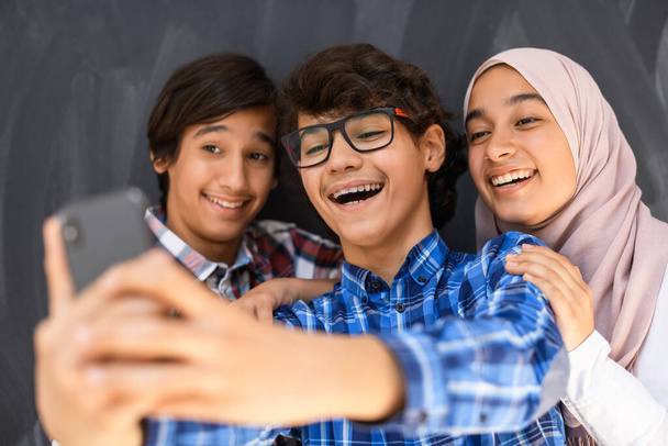 Group of arab teens taking selfie photo on smart phone with black chalkboard in background. Selective focus  - Photo, image