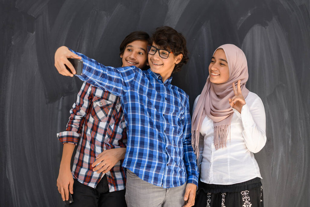 Group of arab teens taking selfie photo on smart phone with black chalkboard in background. Selective focus  - Φωτογραφία, εικόνα
