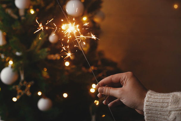 Käsi kädessä polttava kipinä taustalla joulukuusi valot juhlava ilta huoneessa. Hyvää uutta vuotta! Ilotulitus bengal hehkuu naisen kädessä. Tekstiavaruus. Ilmakehän momentti - Valokuva, kuva