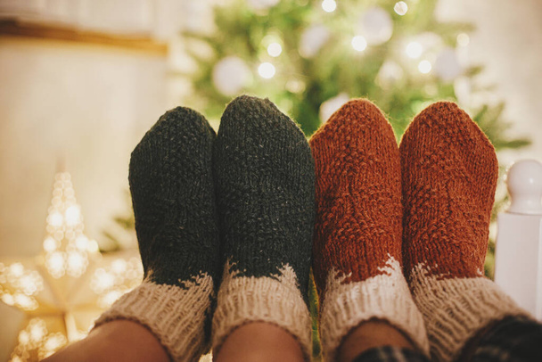 Couples pieds en chaussettes de laine confortables sur le fond de l'arbre de Noël dans les lumières dans la salle de soirée festive. Célébrer des vacances d'hiver ensemble, des moments de famille confortables à la maison. Chaussettes chaudes élégantes - Photo, image