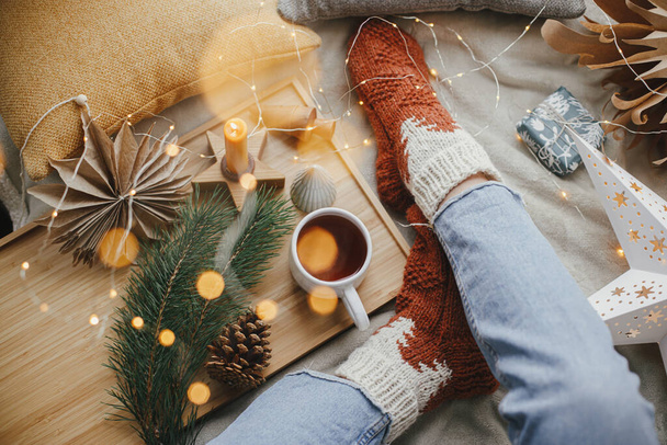 Des moments d'hiver confortables à la maison. Pieds de femme en chaussettes de laine sur lit doux avec tasse de thé chaude, étoiles de Noël, lumières dorées, arbres, bougie et oreillers. Fille se détendre dans la salle de soirée scandinave - Photo, image