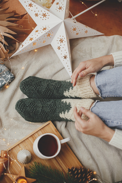 Woman legs in warm socks on soft blanket with cup of tea, christmas stars, golden lights, trees, candle and pillows, top view. Cozy relaxing moments at home. Winter and autumn holidays. - Photo, Image