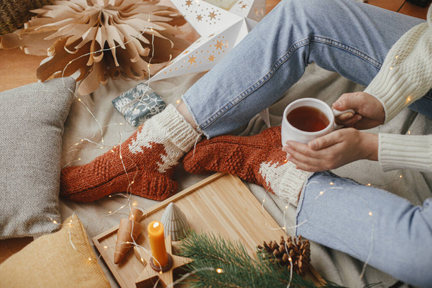 Žena ruce s teplým šálkem čaje relaxaci na měkké posteli s vánoční hvězdy, zlatá světla, stromy, svíčky a polštáře ve skandinávském pokoji. Útulné chvíle doma. Zimní dovolená - Fotografie, Obrázek