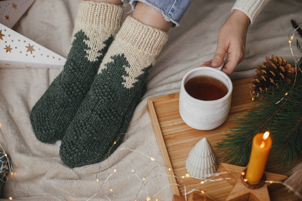 Kellemes téli pillanatok otthon. Női lábak gyapjú zokniban puha ágyban meleg csésze teával, karácsonyi csillagokkal, arany fényekkel, fákkal, gyertyával. Lány pihentető skandináv esti szobában - Fotó, kép