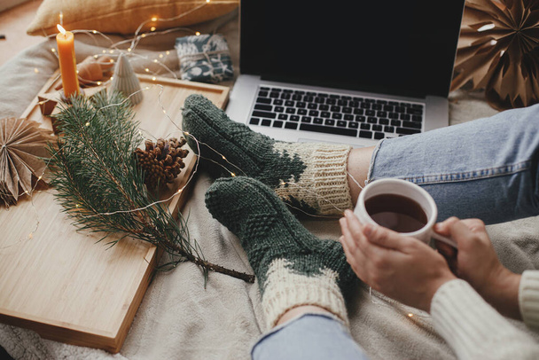 Woman with warm cup of tea relaxing on soft bed with laptop, christmas lights, stars, pine, candle and pillows in stylish room. Cozy winter holidays at home. Freelance and blog - Foto, Imagen