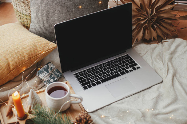 Laptop on soft bed with cup of tea, christmas lights, stars, pine, candle and pillows in scandinavian room. Freelance and blog. Remote work and online. Cozy winter holidays at home. Empty screen - Фото, зображення