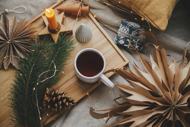 Warm cup of tea with christmas lights, stars, pine trees, candle and pillows on soft bed. Flat lay. Cozy home. Winter hygge. Festive holiday decorations in scandinavian room. Atmospheric  moment - Photo, Image