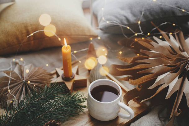 Lämmin kuppi teetä pehmeällä sängyllä joulutähtien, kultaisten valojen, mäntyjen, kynttilän ja tyynyjen kanssa skandaalihuoneessa. Kodikas koti. Ilmakehän maaginen hetki. Hygge talvilomat - Valokuva, kuva