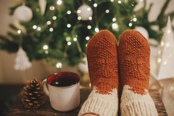 Frauenfüße in kuscheligen Wollsocken und eine Tasse warmen Tee auf dem Hintergrund des Weihnachtsbaums in Lichtern im festlichen Abendzimmer. Gemütliche Wintermomente zu Hause. Stilvolle warme Socken auf weiblichen Beinen - Foto, Bild