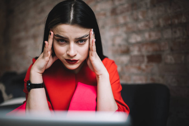 Young good looking lady in red dress with flawless makeup pressing temples and thinking hard while working on serious project on laptop in modern workspace - Photo, image