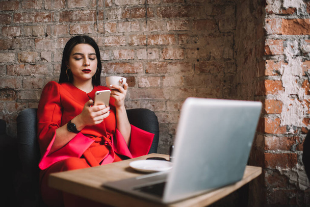 Kırmızı elbiseli iş kadını cep telefonuyla internette geziniyor ve iş sırasında dizüstü bilgisayarla masada otururken mesajları kontrol ediyor. - Fotoğraf, Görsel