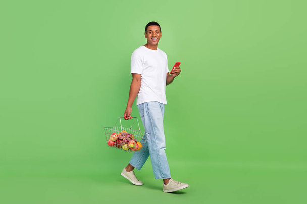 Full length profile side photo of young african man walk shop online use smartphone purchase isolated over green color background. - Valokuva, kuva
