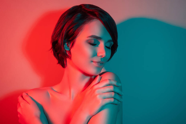 Fénykép nyugodt vidám álmodozó lenyűgöző rövid frizura nő ölelés magát elszigetelt piros neon fény színes háttér - Fotó, kép