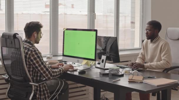 Puukotus, jossa kaksi nuorta monietnistä ohjelmistoinsinööriä kirjoittaa näppäimistöille, katselee tietokonenäyttöjä, istuu työpöydillä modernissa toimistossa - Materiaali, video