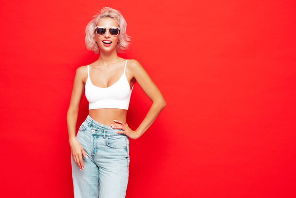 Young beautiful smiling female in trendy summer clothes. Sexy carefree woman posing near red wall in studio. Positive blond model having fun and going crazy. Cheerful and happy. In sunglasses - Foto, afbeelding