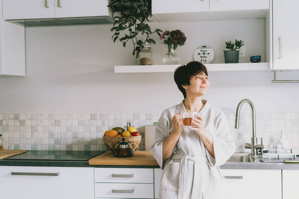 Happy woman in the bathrobe holding a cup of fresh tea in her modern white kitchen. Good morning concept. Positive mood. Selective focus. Copy space. - Photo, image