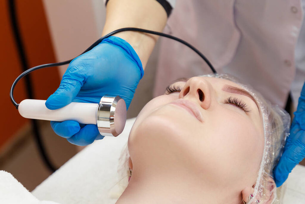 The process of cavitation on the face of a young woman. Rejuvenation and tightening of the skin of the face of hardware cosmetology. The hands of the beautician holds the handpiece of radio frequency cavitation near the woman's face. - Photo, image