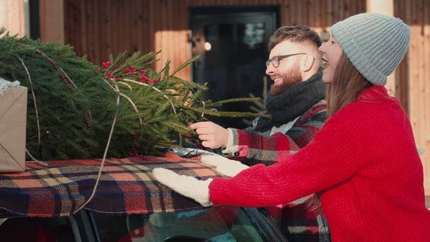 Young beautiful woman in red sweater puts Christmas tree on car roof with happy friends at winter house slow motion. - Foto, immagini