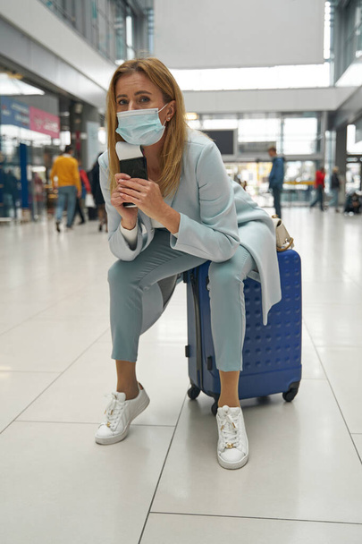 Low-spirited tourist in face mask sitting on her baggage - 写真・画像