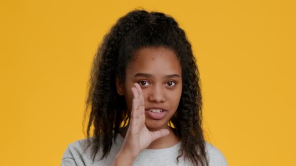 Musta tyttö kuiskaa salaisuuksia elehtiminen sormi huulilla, keltainen tausta - Materiaali, video