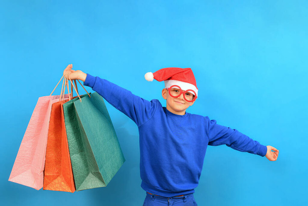 Ein fröhlicher Junge mit Nikolausmütze hält Pakete mit Geschenken und winkt mit den Händen zur Seite. - Foto, Bild