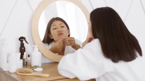 Problema dermatologico. Giovane infelice asiatico signora squeezing brufoli su suo mento, guardando specchio in bagno - Filmati, video
