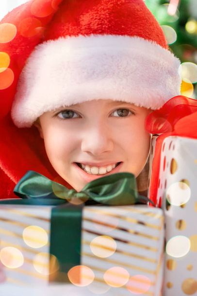 Happy boy in a Santa hat looking at camera with many gifts boxes at morning at home. Merry Christmas concept. Smiling joyful child in a winter holiday. Vertical portrait with bokeh lights. - Foto, Imagen